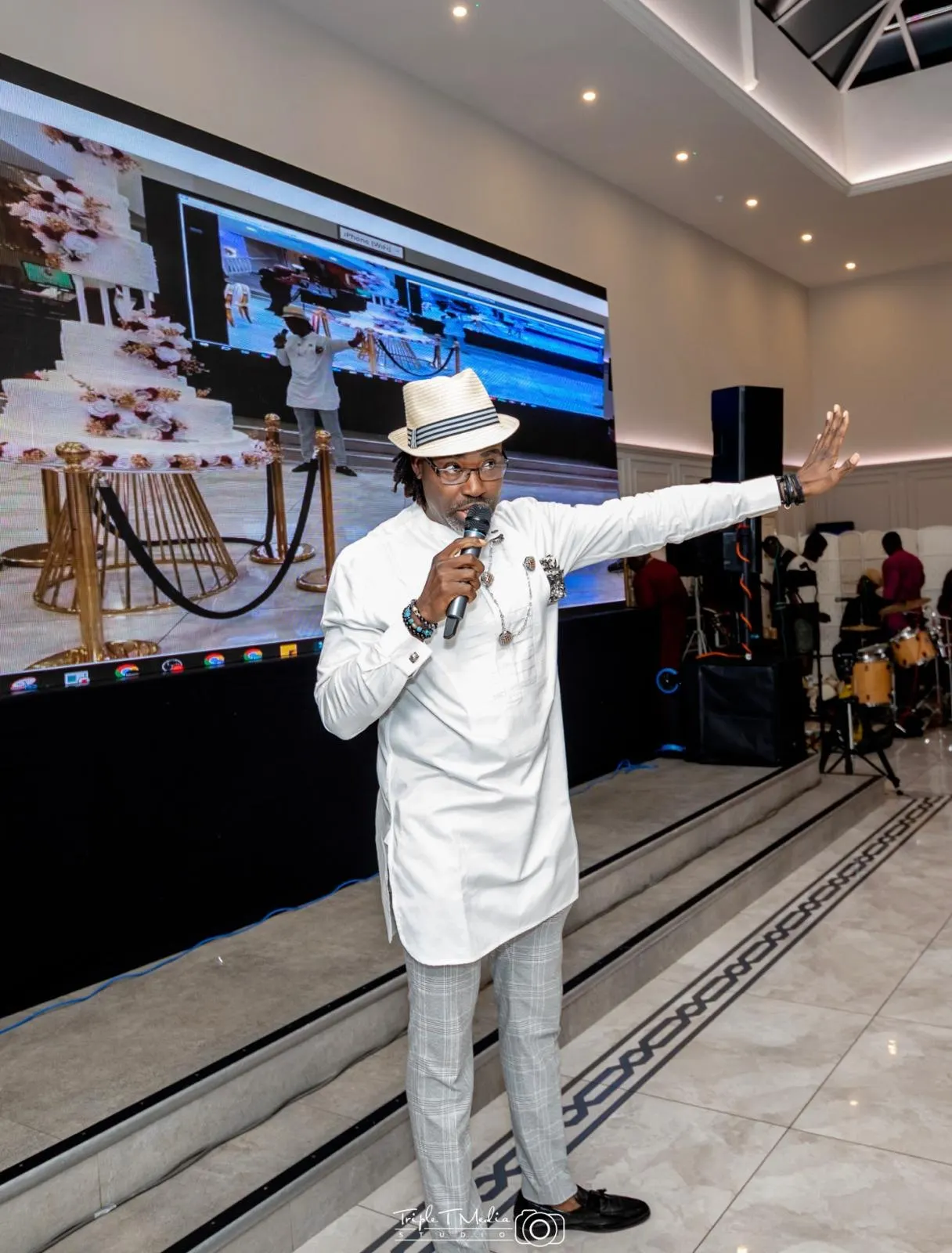 A host in white traditional attire and a straw hat speaks into a microphone in front of a large screen