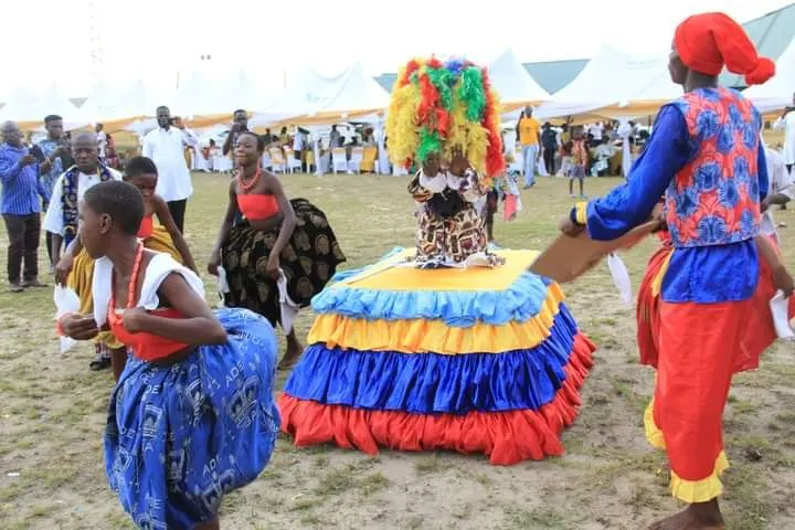 A masquerade in a tiered red, blue and yellow costume, with dancers in wrappers around it